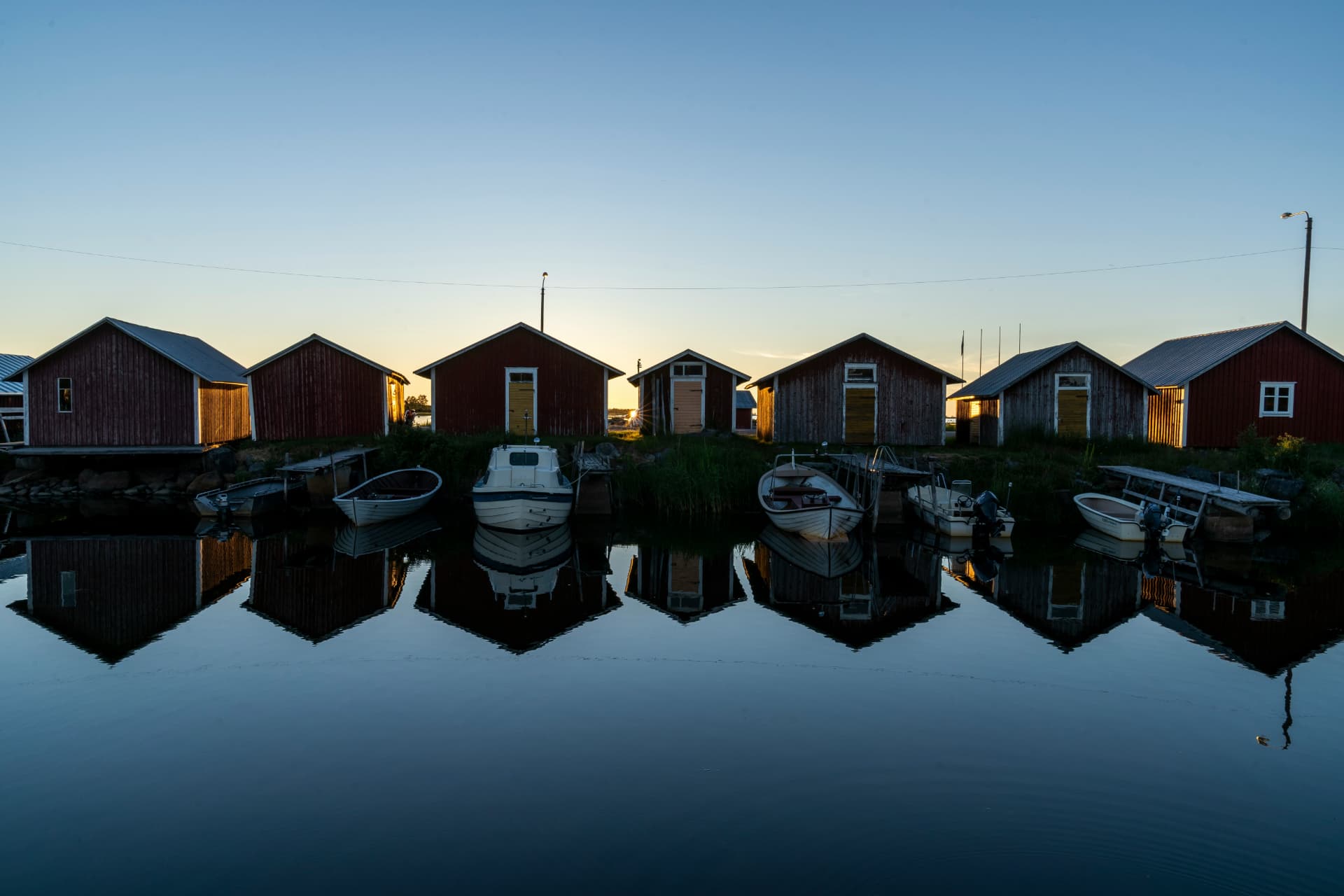 Red boathouses and moored boats mirrored in still archipelago water as the sun sits on the horizon