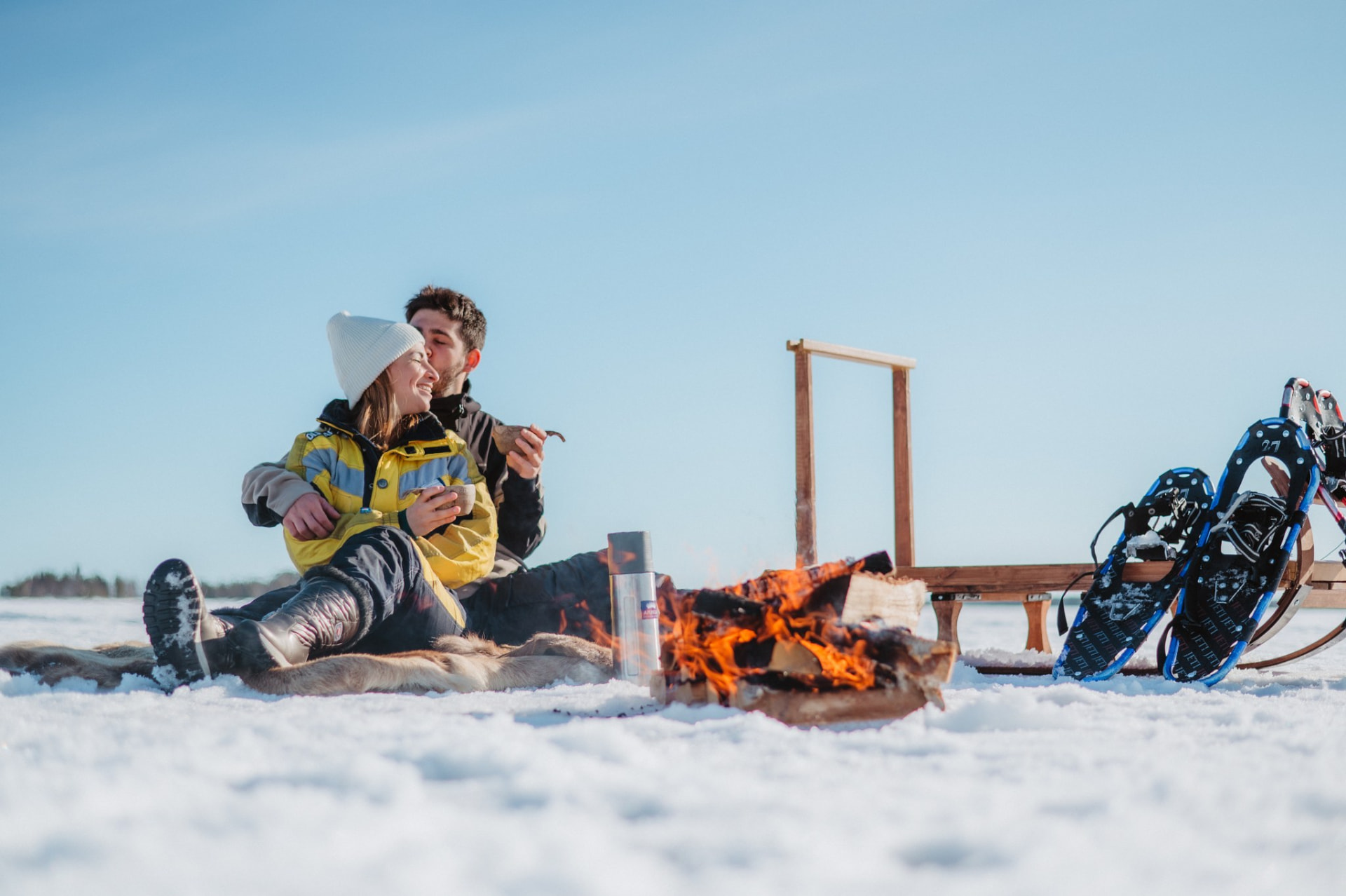 Guests resting on reindeer hides by an open fire on a frozen lake, snowshoes beside them